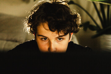 Teen boy with curly hair focused on computer screen, illuminated by warm indoor lighting
