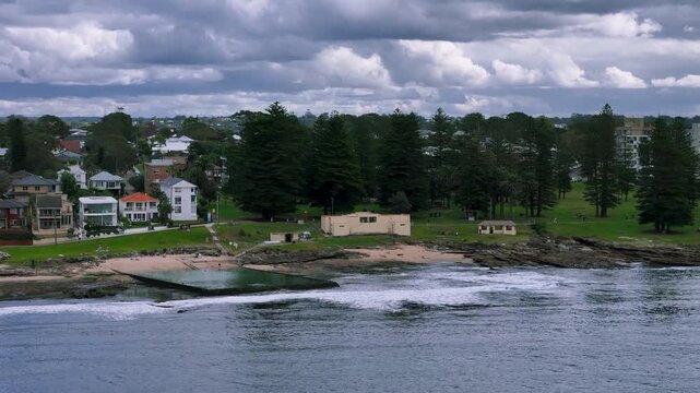 Cronulla, Sheily Beach &ndash;UHD Drone Video- Flying from the sea above the surf towards the Ocean Pool and Sheily Park with the large Norfolk Pine trees on the beach, typical for Sydney, Australia