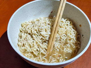 Chewy white instant noodles are held up in a white bowl, highlighting their texture and deliciousness, against the blurry background of the bowl.