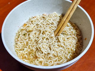 Chewy white instant noodles are held up in a white bowl, highlighting their texture and deliciousness, against the blurry background of the bowl.