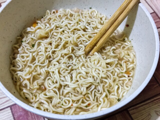 Chewy white instant noodles are held up in a white bowl, highlighting their texture and deliciousness, against the blurry background of the bowl.