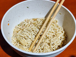 Chewy white instant noodles are held up in a white bowl, highlighting their texture and deliciousness, against the blurry background of the bowl.
