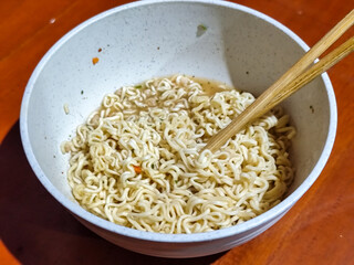 Chewy white instant noodles are held up in a white bowl, highlighting their texture and deliciousness, against the blurry background of the bowl.