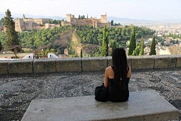 Fototapeta premium Tourist woman sitting on a stone bench and admiring the panoramic view of the Alhambra palace and fortress in Granada from Mirador de San Nicolas Andalusia. Spain