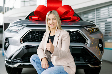 Happy businesswoman showing thumbs up near new car with red bow in dealership