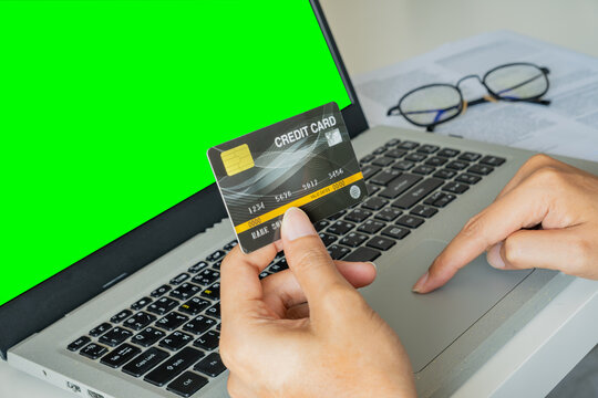 A person holds a credit card while browsing on a green-screen laptop—signaling online shopping. Nearby, glasses and documents hint at a casual work-from-home setup. Green screen for copy space.