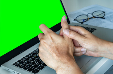 An Asian man holds his thumb, showing signs of pain or strain. He sits at a desk near a laptop,...