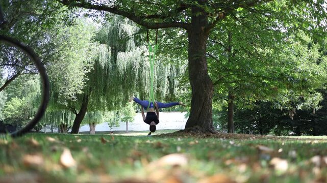Woman performs an aerial yoga inversion suspended from a tree in a tranquil green park surrounded by weeping willows and sunlight creating a peaceful and meditative scene