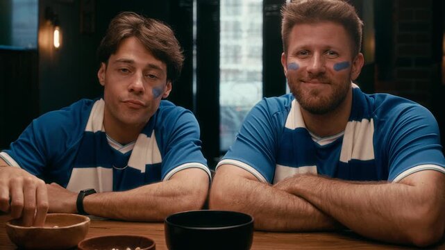 Two guys in blue t-shirts and striped scarves sitting by table in bar during broadcast of football match, looking at each other, then turning to camera while one of them eating snacks