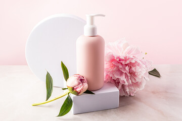 Pink unbranded plastic dispenser pump bottle standing on a white square podium with peony. packaging mockup. bottle of liquid soap, gel, shampoo.
