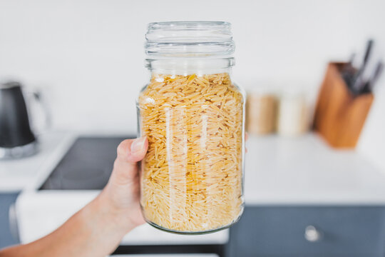 hand holding glass jar of risoni wheat pasta in front of kitchen bokeh, cooking from scratch and having an organised pantry