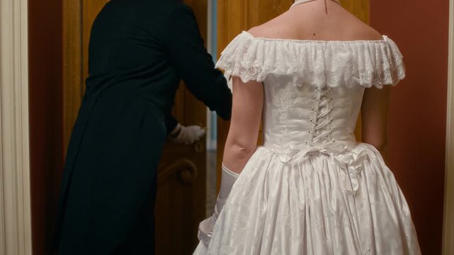 Back view cropped shot of graceful noblewoman wearing elegant white dress and gloves entering grand ballroom as servant opening wooden doors for her