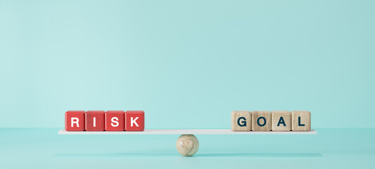Wooden blocks spelling “RISK” and “GOAL” balanced on seesaw plank with sphere base. Concept of risk management, business strategy, investment decision, life goals.