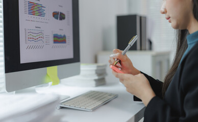 Businesswoman reviewing charts and graphs on a computer monitor, taking notes with a pen, focused...