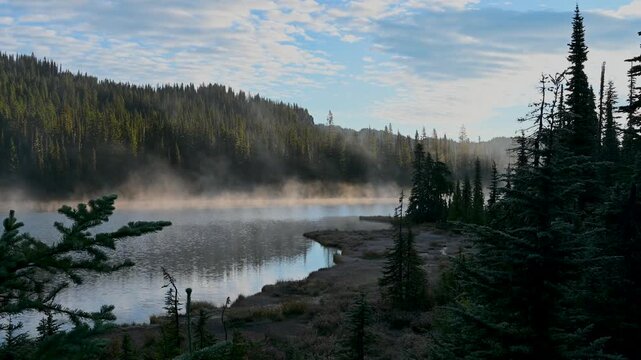 Aerial view of mist rising from a tranquil alpine lake bordered by dense evergreen forest.