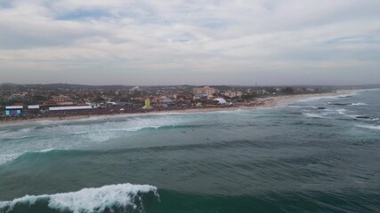 Drone captures the 2025 WSL surf event at Itaúna Beach, Saquarema, Rio de Janeiro. Huge crowd, strong waves, surfers, tents and big screens under dramatic cloudy skies by the Atlantic Ocean.