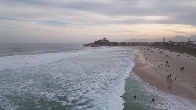 Drone view of Ita&uacute;na Beach in Saquarema, Rio de Janeiro, known for hosting the World Surf League (WSL). Sunset scene with calm waves, beachgoers and the iconic church on the hill by the coast.