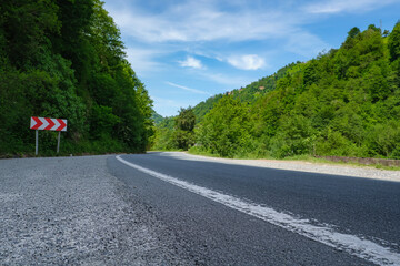 Winding asphalt road through forest with traffic sign. View of a winding road bordered by lush greenery and trees with blue sky with fluffy clouds. idea for exploration and travel.