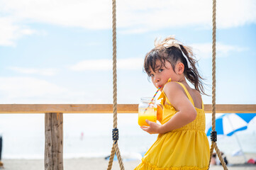 Japanese girl in a yellow dress relaxing on a seaside swing while holding a mason jar with orange...