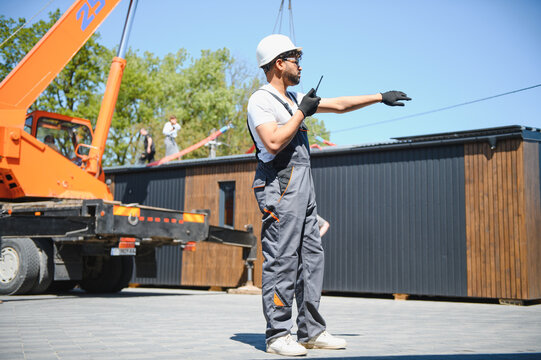 Construction worker directing crane lifting modular house onto truck