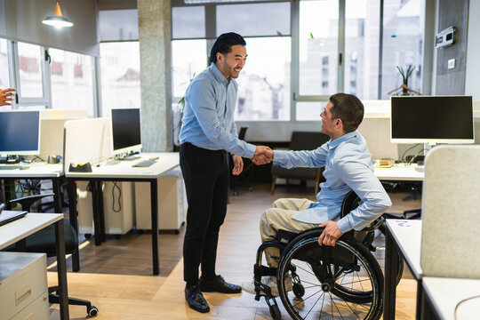 Two businessmen shaking hands in a modern coworking office, showing respect and collaboration in an inclusive work environment, one of them is using a wheelchair and the other is chinese