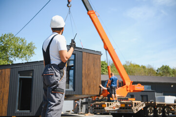 Construction worker supervising modular house lifting onto truck by crane