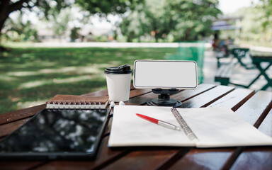 mobile phone on stand with coffee cup, digital tablet and notebook on wooden table in park, working outside concept and relax via modern technology from mobile