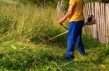 A man is trimming tall grass with a gas-powered weed trimmer near an wooden fence in backyard, overgrown outdoor area. Trees and plants surround the scene and sunlight highlights the natural setting