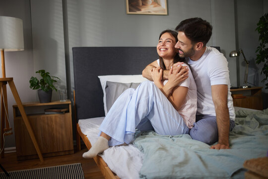 A joyful couple sits on a neatly made bed in a modern bedroom, embracing while laughing and enjoying each other's company on a relaxed morning.