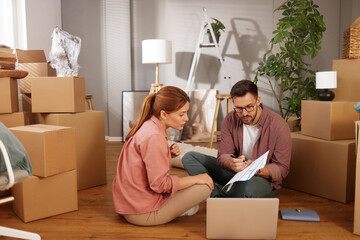 Two individuals sit on the floor discussing their upcoming move, surrounded by moving boxes and...