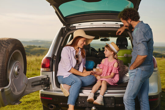 A family gathers for a picnic on a warm afternoon, sharing snacks and laughter while seated at the back of their SUV. The setting features grassy hills and a clear sky, creating a joyful atmosphere.