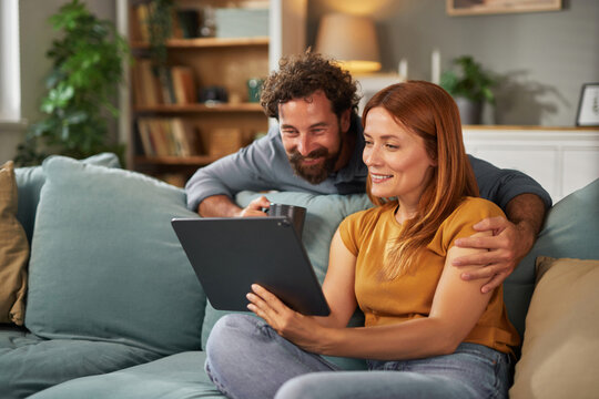 A couple is seated on a comfortable couch, sharing a joyful moment as they watch something entertaining on their tablet. They appear relaxed and engaged in conversation, surrounded by homey decor.
