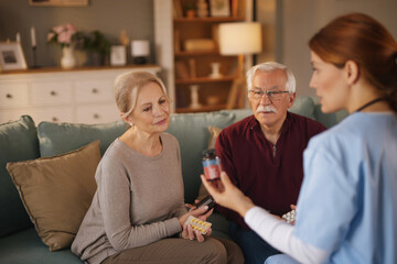 A home health nurse in blue scrubs discusses medication instructions with an elderly couple.