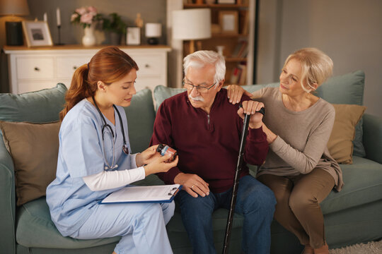A healthcare worker in scrubs shows a pill bottle to an elderly man holding a cane as his wife sits beside him with her hand on his shoulder in the bright living room of his home.