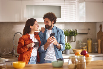 A couple stands together in their home office, smiling and looking at a smartphone. They are...