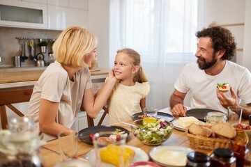 A mother, father, and daughter are seated at a dining table filled with various dishes, laughing...