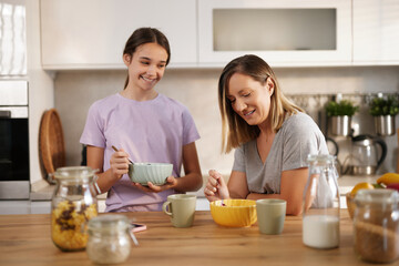 A mother and daughter enjoy breakfast together in a sunny kitchen, with bowls of cereal and cups of coffee. Laughter and warmth fill the air as they bond over their meal.