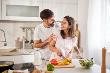 In a well-lit kitchen, a couple shares a joyful moment while cooking. They laugh and interact as they chop vibrant vegetables for a healthy meal, surrounded by fresh ingredients.
