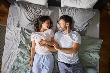 Two people are lying side by side on a bed full of pillows and blankets, smiling and holding hands, creating an intimate and cheerful connection during the morning.