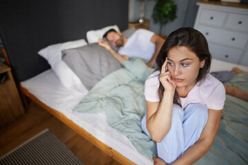 A woman sits on the edge of the bed, looking concerned while a man sleeps peacefully beside her....