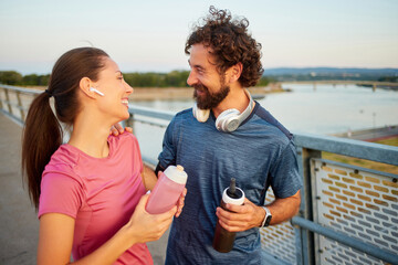 Two joggers take a break on a riverside path, smiling and chatting as they hydrate. The sunset...