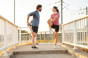 Two friends engage in stretching exercises outdoors, preparing for a morning workout. The sun...