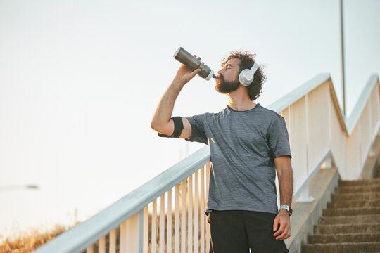 Jogger pauses on outdoor stairs to drink water from a stainless steel bottle while wearing headphones, emphasizing fitness during early evening exercise routine. - Powered by Adobe