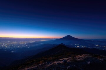 Pre-dawn vista of a majestic volcano, silhouetted against a twilight sky, overlooking a sprawling city lightscape nestled in valleys below