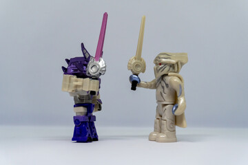 Two toy figures face each other, one purple and one tan, each holding a lightsaber, in a duel pose...