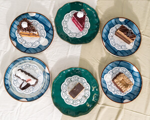 Six assorted cake slices on patterned plates, arranged on a light fabric surface, shot in flat lay composition with natural lighting.