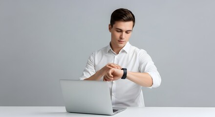 A man in a white shirt checking the time on his smartwatch in a studio setting with a laptop computer on a table. The background is plain gray.
