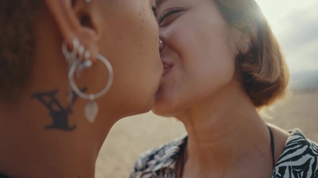 Passionate lesbian couple sharing tender moment while embracing and kissing on sandy beach during romantic sunset, expressing deep love and connection