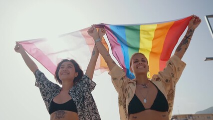 Female couple running along sandy coastline, passionately waving rainbow flag, celebrating love, freedom, and lgbtq+ pride during sunny summer vacation - Powered by Adobe