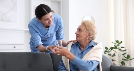 Fototapeta premium Nurse giving glass of water to senior woman indoors. Home health care service